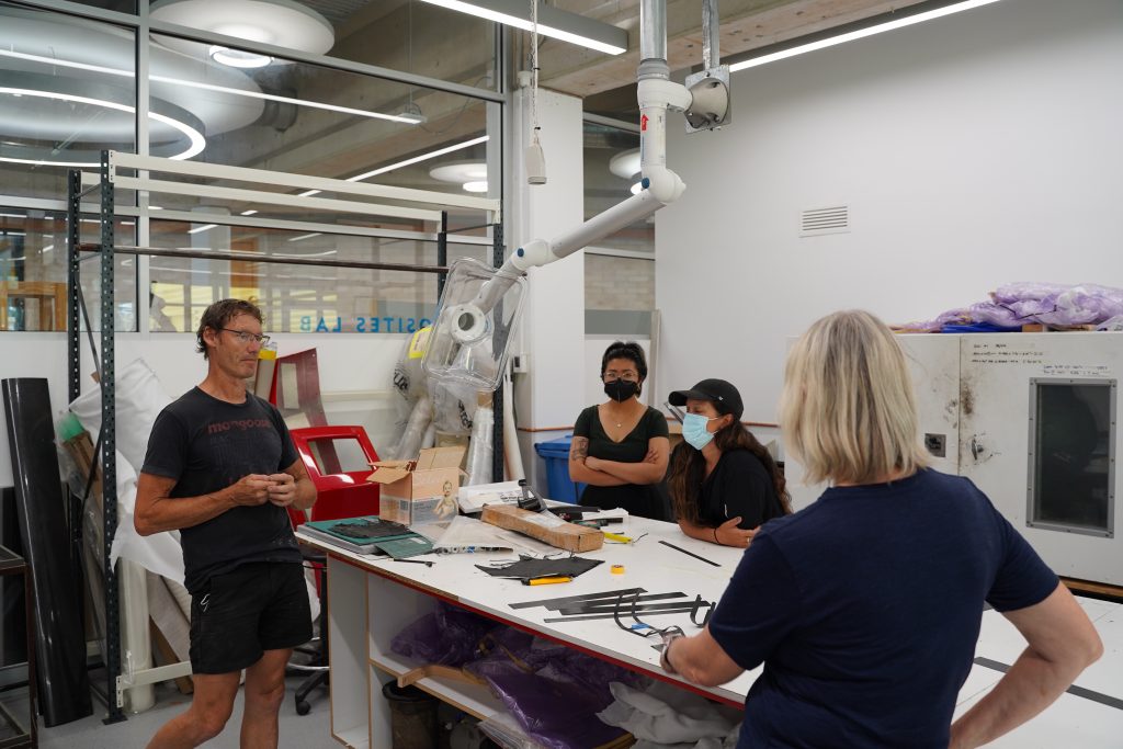 Three artists (second left to right: Zhi, Melanie Lane, Kirsten Wehner) are talking to Bill the technician (first left) at UC Workshop 7 about fibre fabric. The space featured in the background shows big machines and a long workbench in the centre, with some black rectangular pieces of fibre fabric on the bench. 